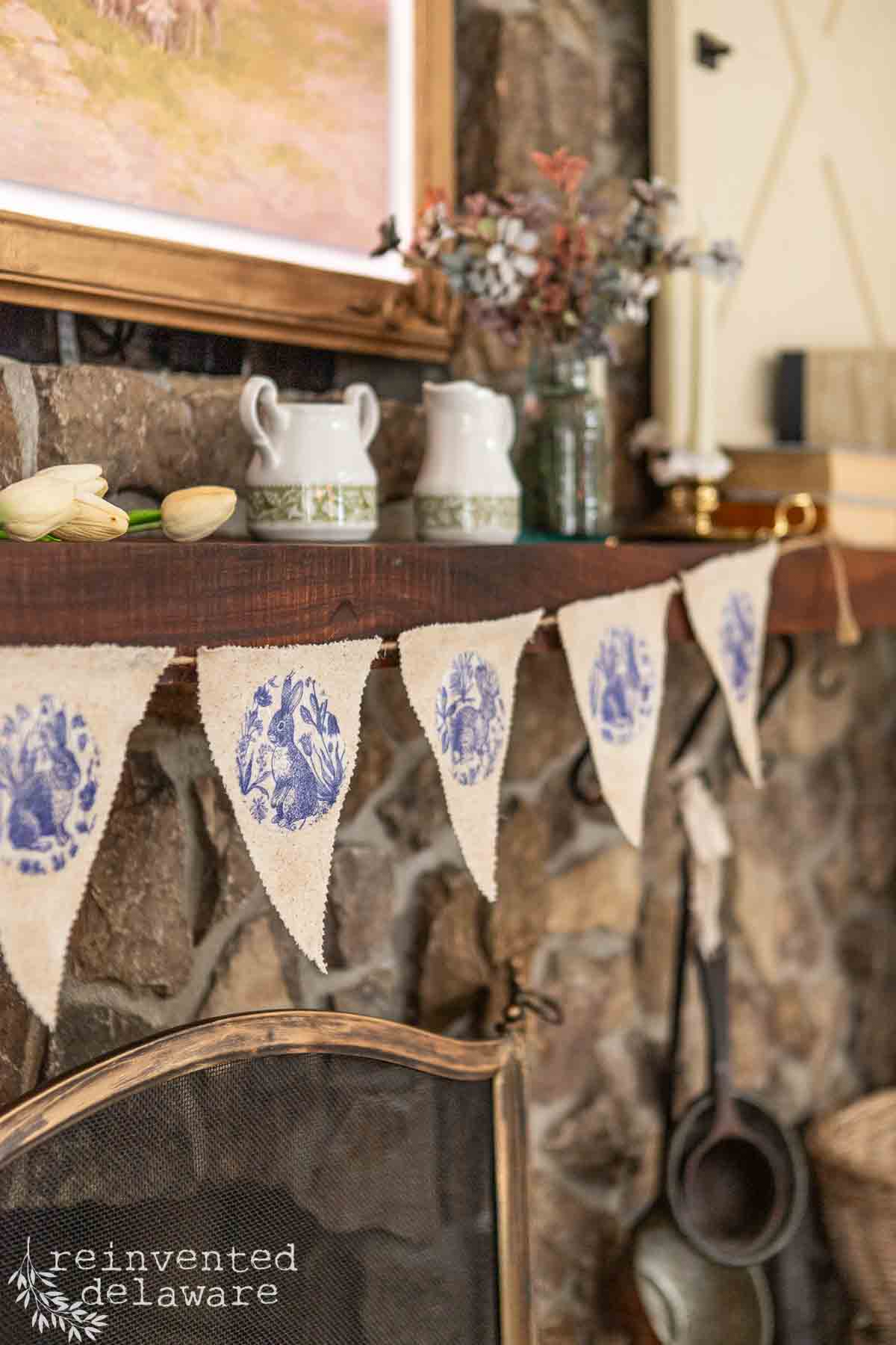 A rustic stone fireplace adorned with a DIY Easter garland of cream fabric flags featuring blue designs. The wooden mantel holds vintage items like white pitchers, a glass jar, and stacked books. Below, a metal fireplace screen adds to the charm.