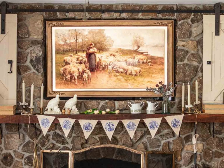 A rustic stone fireplace adorned with candles, a pair of white dove sculptures, and blue-patterned pottery. Above, a painting depicts a shepherdess with sheep. A DIY Easter garland featuring decorative bunting with blue designs is draped across the mantle.