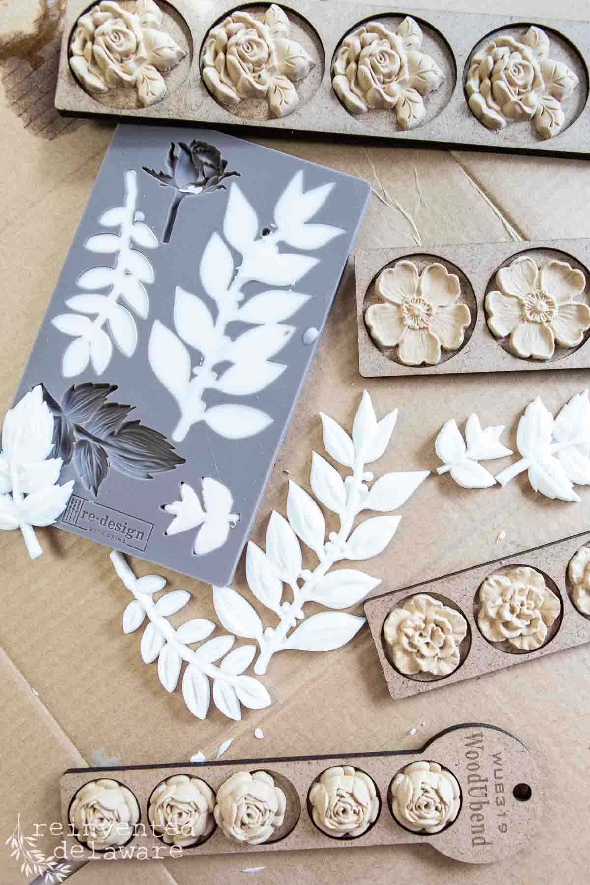 Various floral molds and clay decorations are arranged on a cardboard surface. The molds feature different flower and leaf designs, with some completed pieces in white and beige tones.
