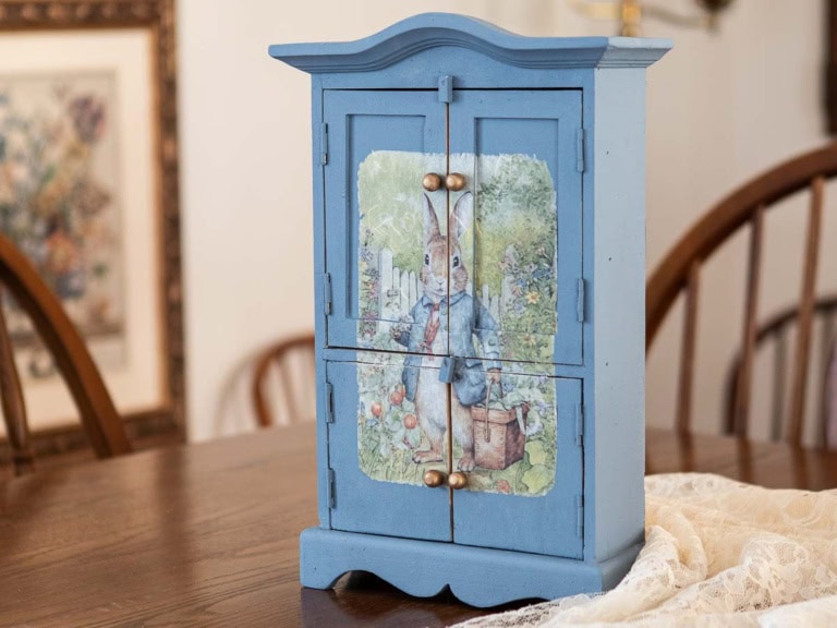 A small, blue wooden cabinet with a painting of a rabbit in a blue jacket on its front. This charming DIY makeover features four doors with brass knobs and sits gracefully on a wooden table, where a lace cloth is partially visible.