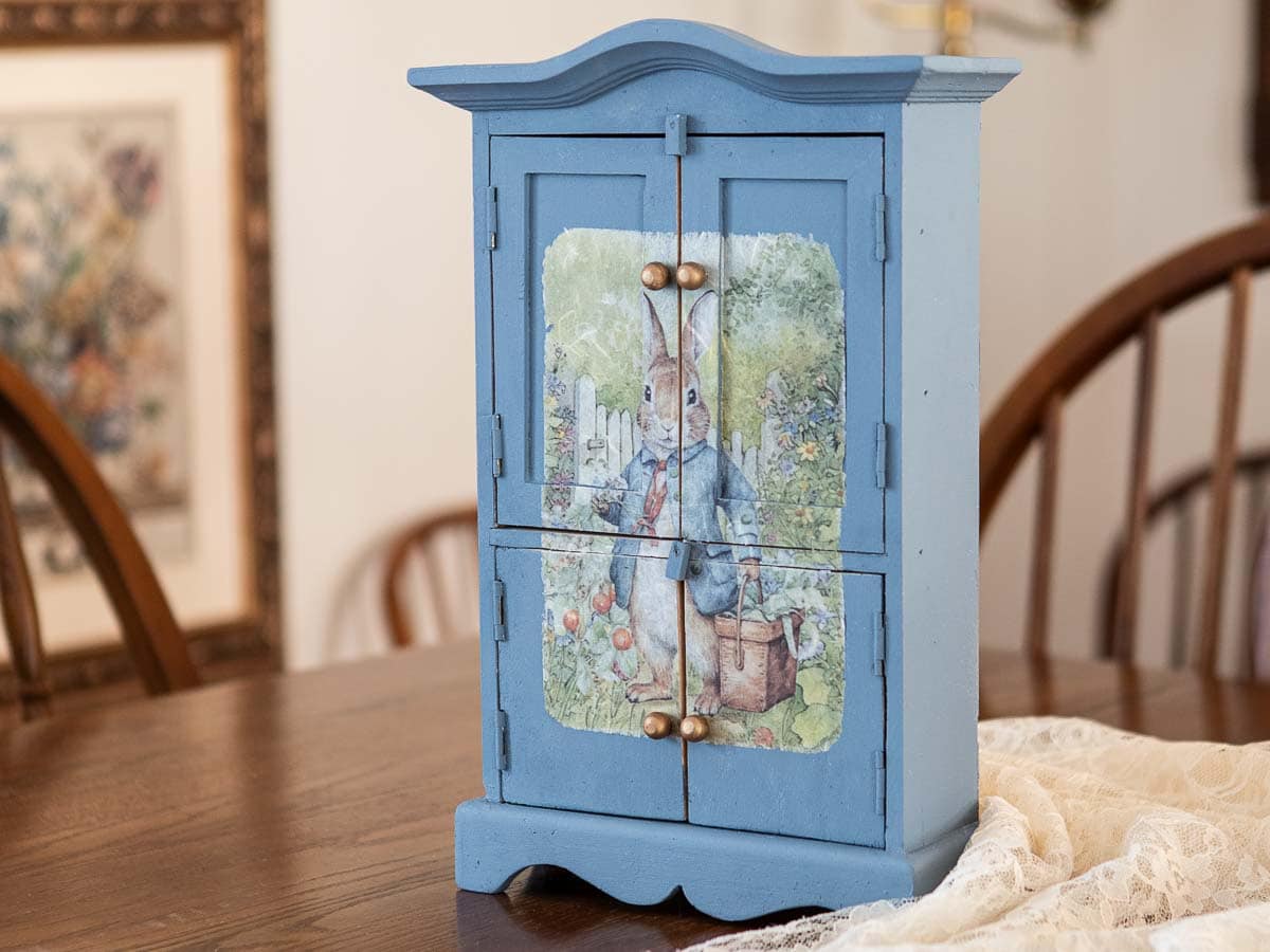 A small, blue wooden cabinet with a painting of a rabbit in a blue jacket on its front. This charming DIY makeover features four doors with brass knobs and sits gracefully on a wooden table, where a lace cloth is partially visible.