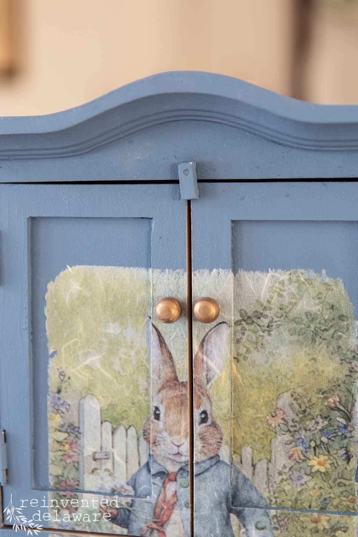 Close-up of a blue cabinet with round golden handles. The doors, part of a DIY makeover, feature a painted image of a rabbit in clothes near a white picket fence with flowers, evoking the charm of a toy. The text "reinvented delaware" is visible at the bottom left.