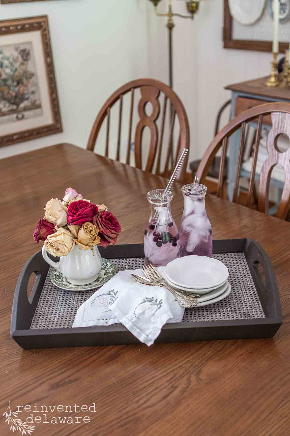 A dark thrift store tray makeover sits on a wooden dining table, holding a small pitcher of faded roses, two glass carafes with berry-infused water, stacked white plates, and embroidered napkins. Wooden chairs and framed art are in the background.