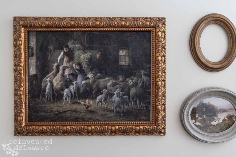 A wall displays a framed painting of two people herding sheep with lambs, alongside two smaller decorative frames—one oval and empty, the other showing geese by water—creating a charming diy vintage wall art display.