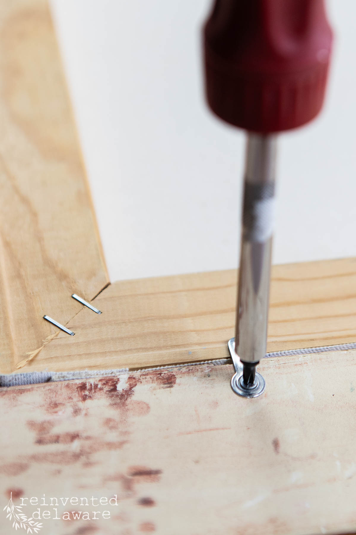 Close-up of a screwdriver tightening a screw into a wooden frame, with staples securing fabric—a perfect diy vintage wall art project. The corner of the frame and distressed surface appear, along with the "reinvented delaware" logo.
