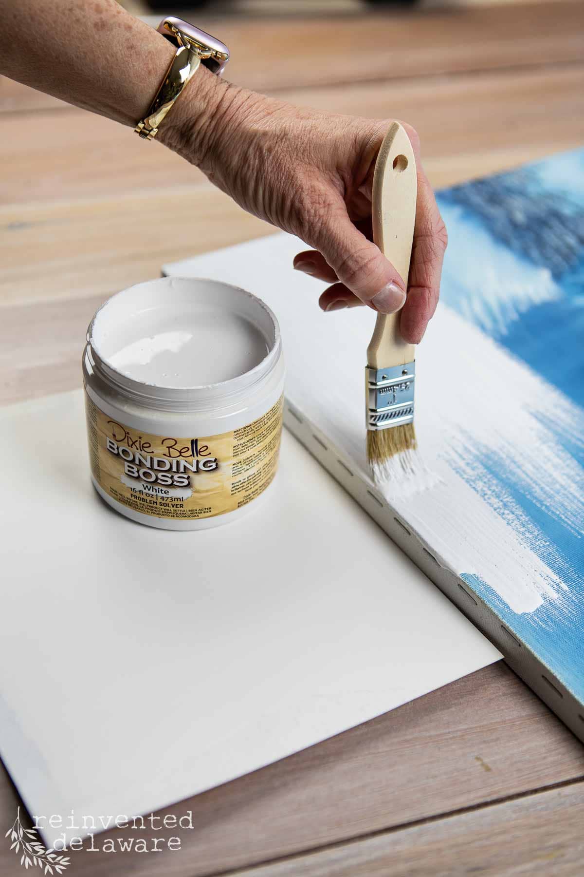 A hand applies white Bonding Boss primer to a canvas with a paintbrush, prepping for diy vintage wall art; an open jar of Dixie Belle Bonding Boss rests on a wooden table nearby.
