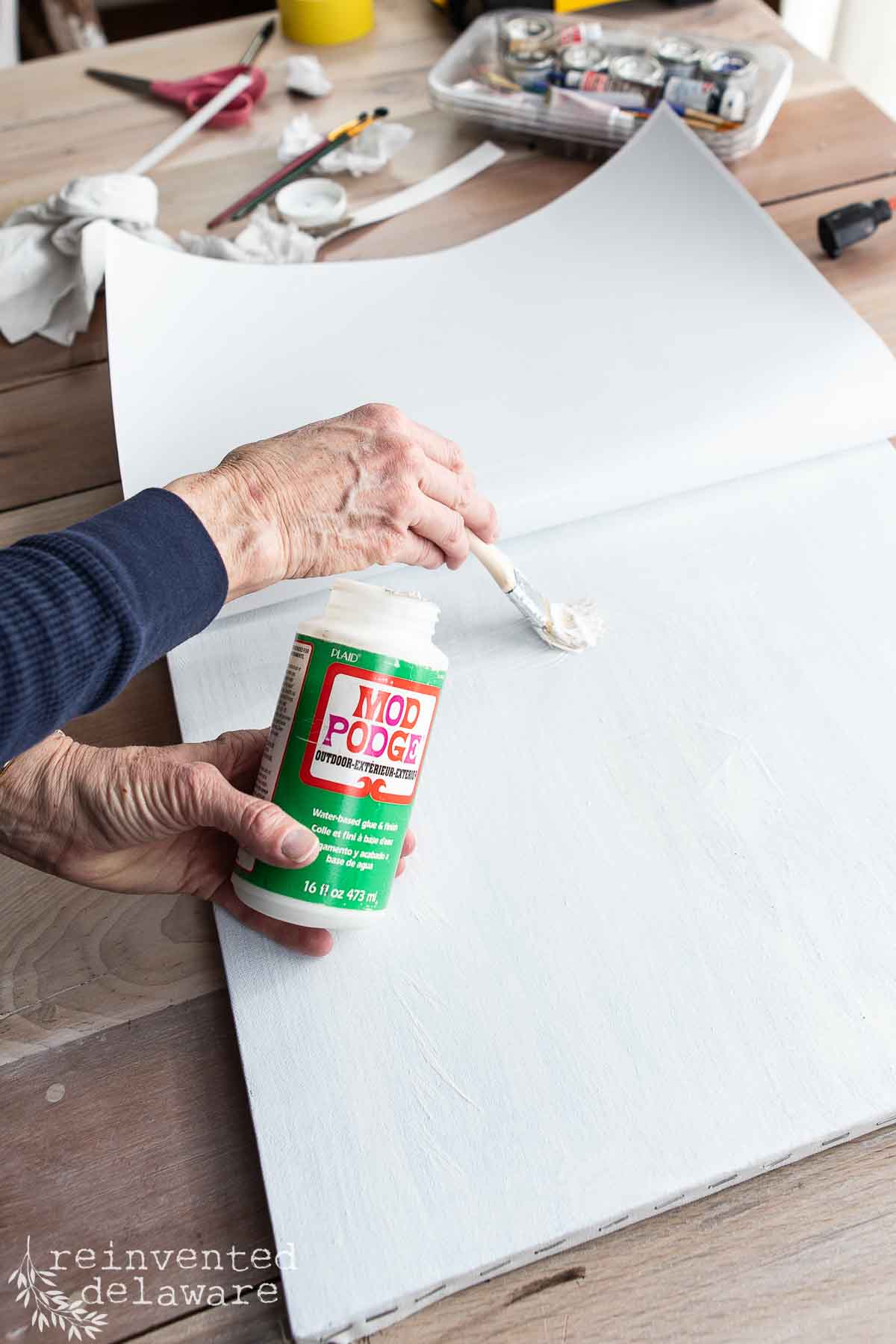 A lady applies Mod Podge to a large white canvas using a paintbrush, creating diy vintage wall art. Art supplies and tools are scattered on the wooden table in the background, setting the scene for a creative crafting project in progress.