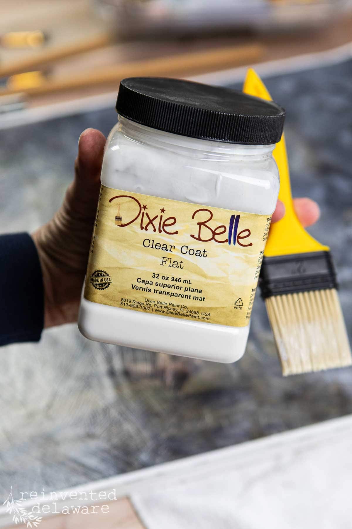 A hand holds a jar of Dixie Belle Clear Coat Flat finish next to a yellow-handled paintbrush. The background is blurred, showing a craft or work surface.