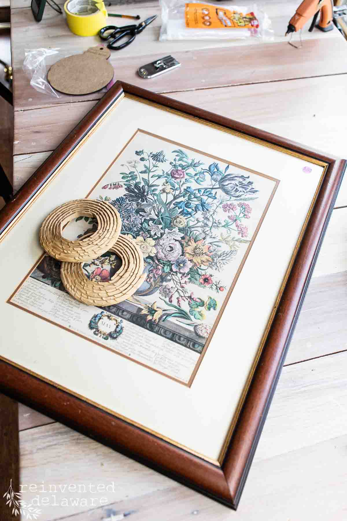 A floral print in a brown wooden frame sits on a table with decorative trim pieces placed on its glass—an inviting example of vintage decor on a budget. Craft supplies like tape, scissors, and glue are scattered around.