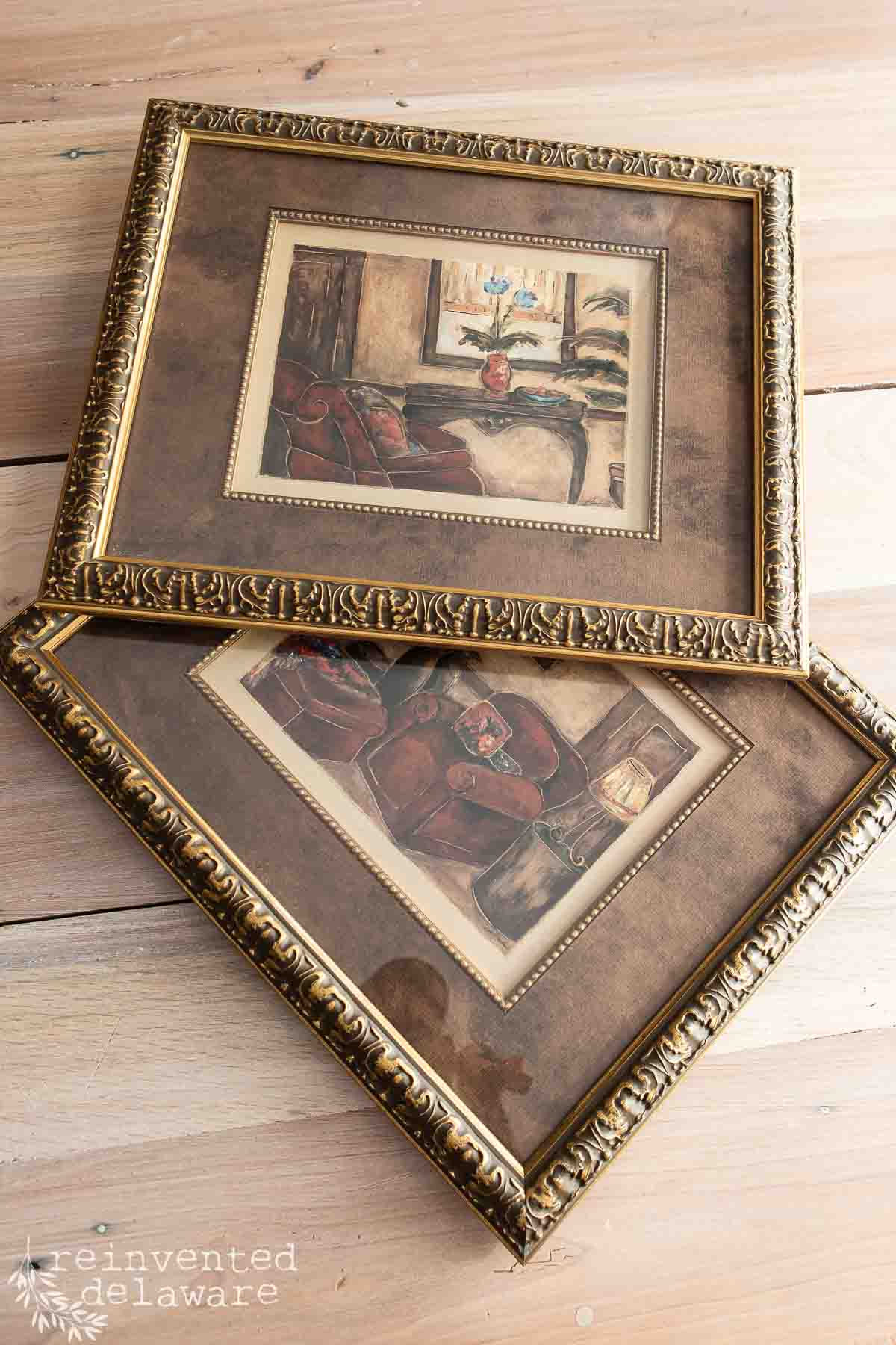 Two vintage-style framed paintings with ornate gold borders, featuring cozy indoor scenes with armchairs, a table, and warm-toned decor, placed on a light wood surface.