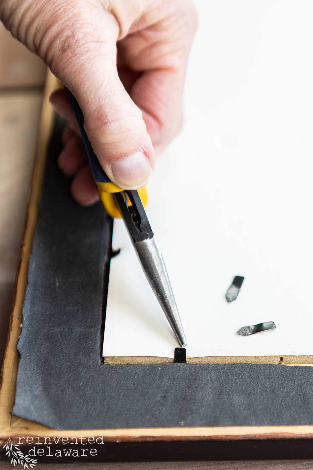 A hand uses pliers to bend a small metal tab on the back of a repurposed picture frame to secure the backing in place. Two loose metal tabs are nearby.