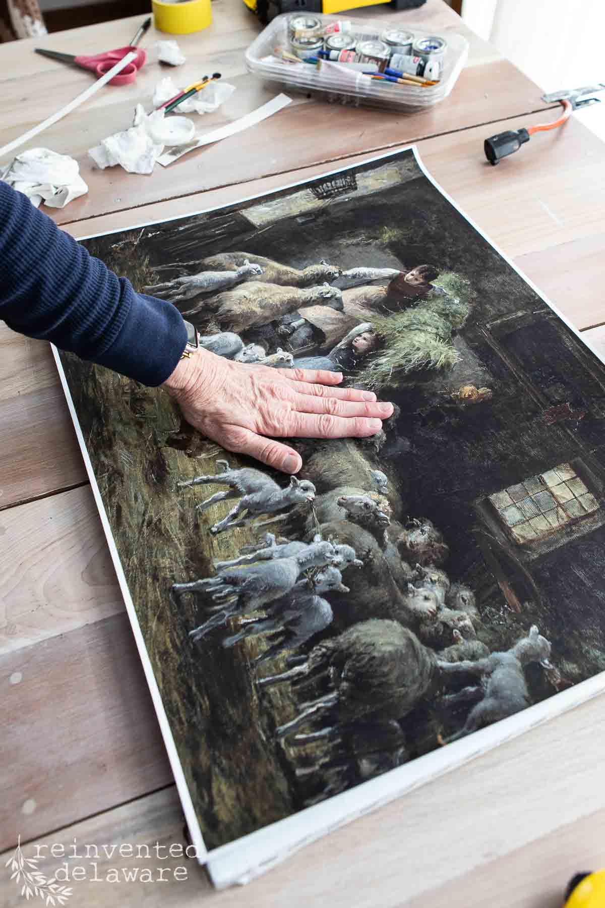A person’s hand smooths a vintage-style painting of sheep and a shepherd on a wooden table, surrounded by art supplies, craft tools, and repurposed picture frames.