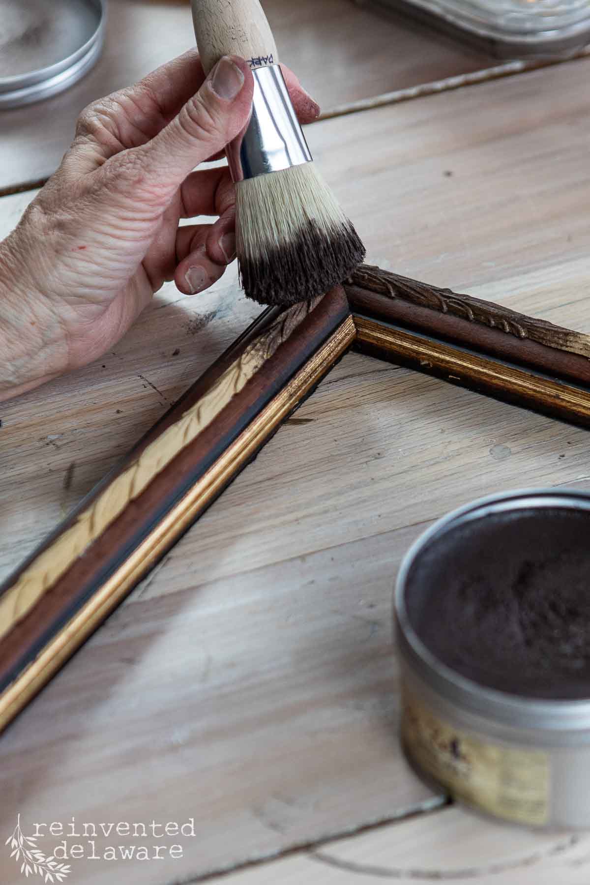 A hand uses a round paintbrush to apply dark wax to the corner of a wooden picture frame on a light wood table, with an open tin of wax nearby—an easy way to revamp old picture frames.