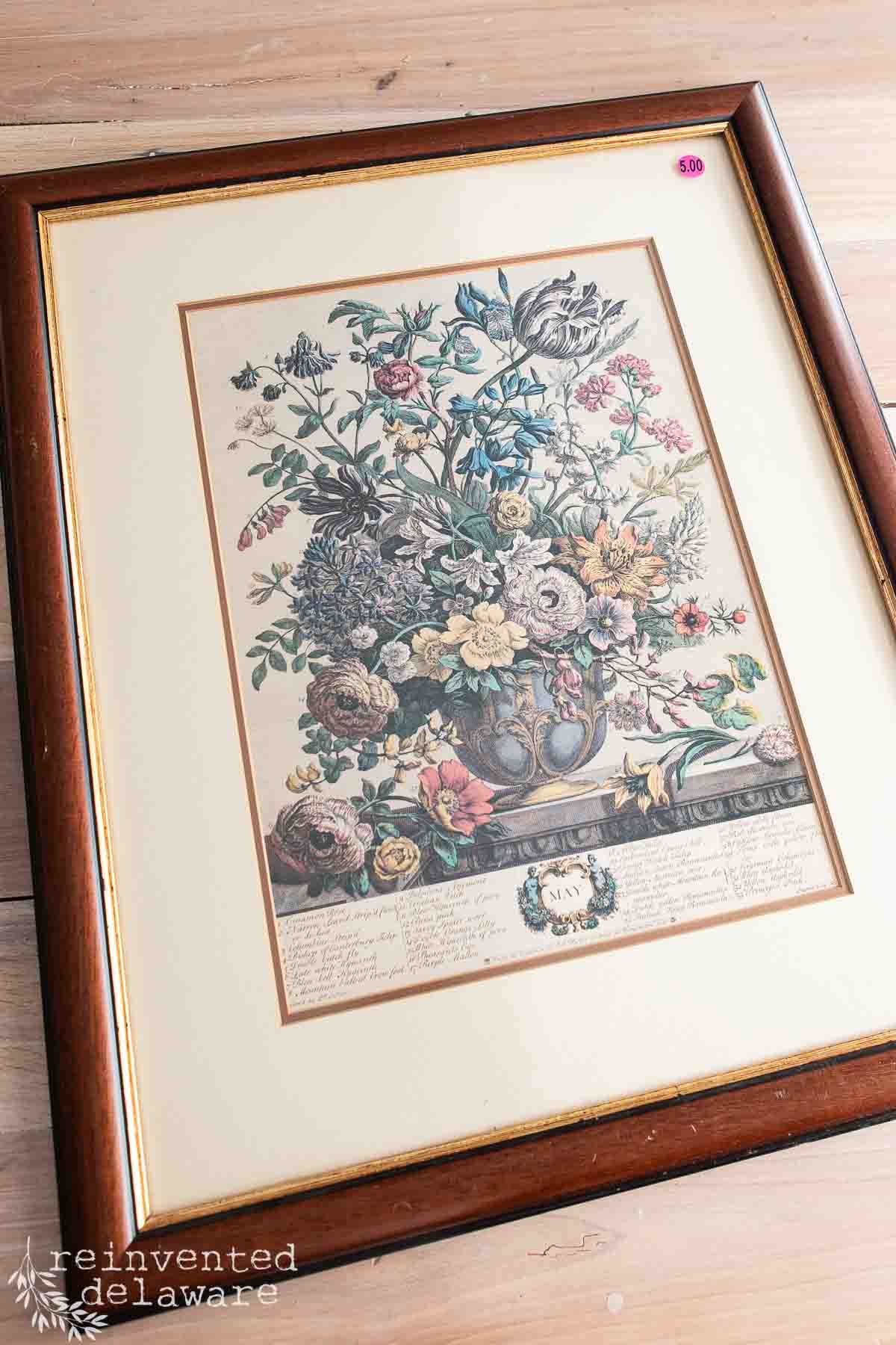 A framed vintage botanical print features a detailed illustration of various colorful flowers in a vase, with descriptive text and labels beneath the image. The frame is dark wood with a cream-colored mat.