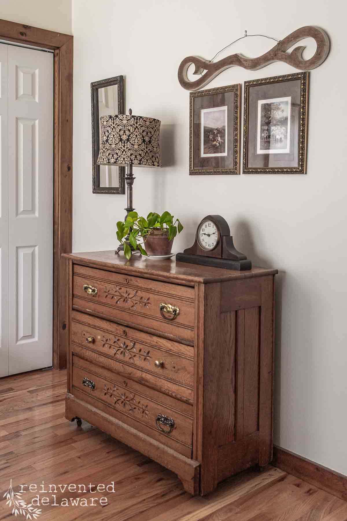 A wooden dresser with brass handles stands against a wall, topped with a lamp, potted plant, and clock. Above it are two framed pictures—one in an upcycled secondhand frame—and a decorative wooden piece. Wood floor and door frame are visible.