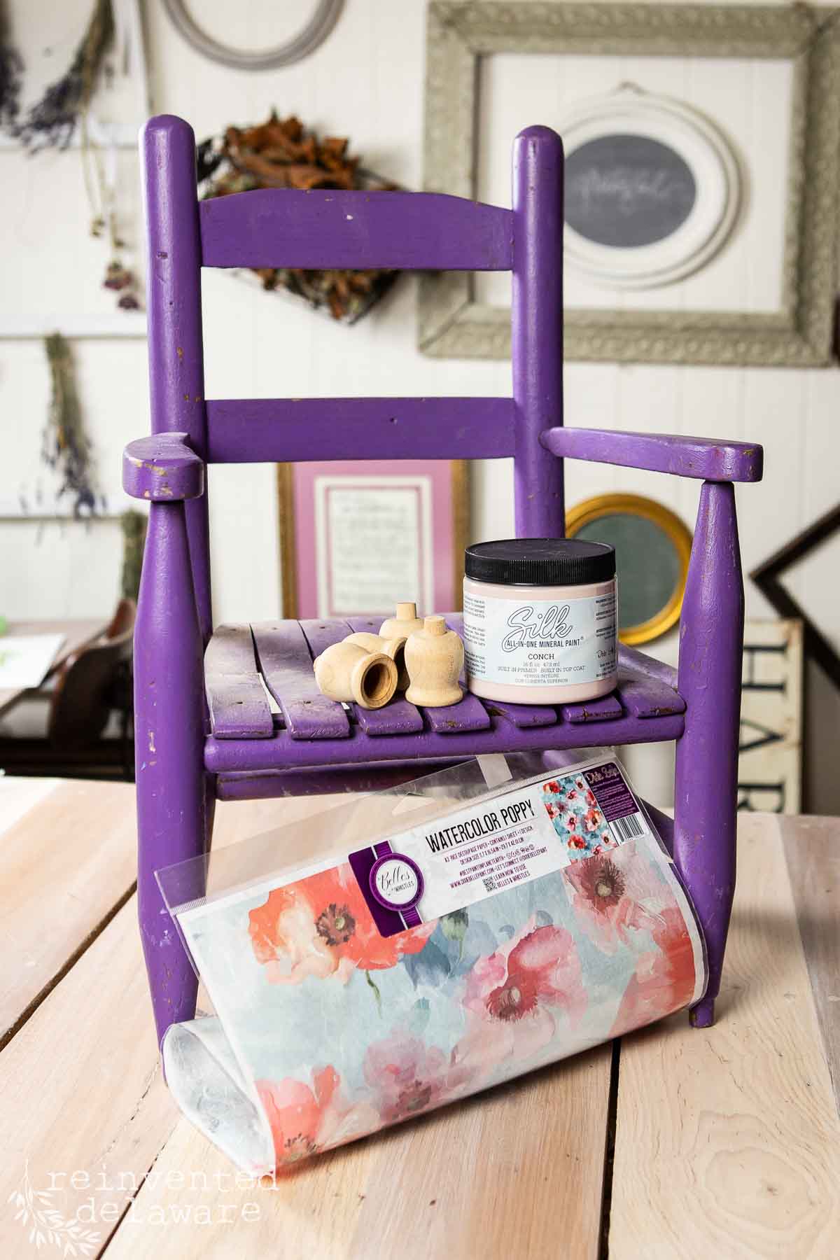 An upcycled chair makeover features a small purple wooden chair on a table, holding a floral transfer sheet, wooden toy parts, and paint, with empty picture frames hanging on the wall in the background.