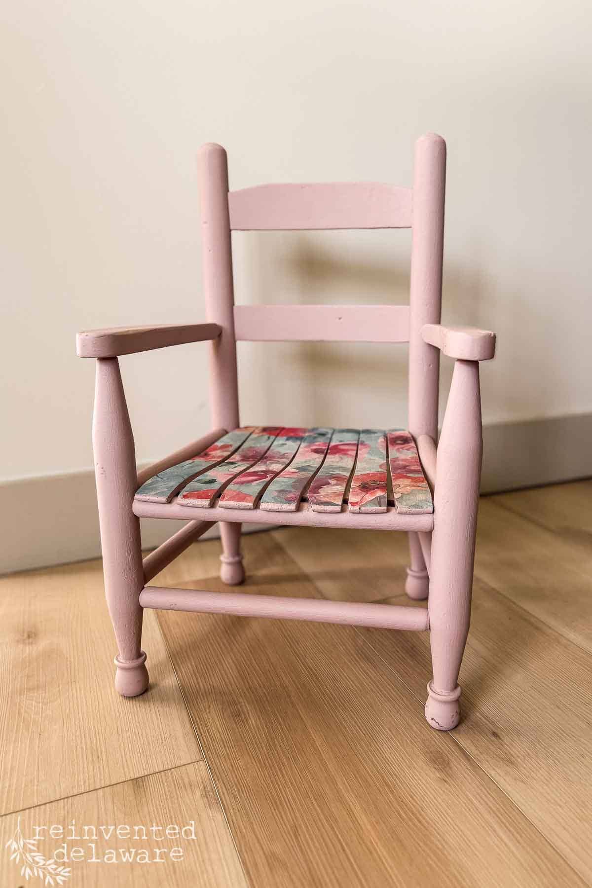 A small pink upcycled chair makeover with floral-patterned slats on the seat sits on a light wood floor against an off-white wall. The chair has armrests and a simple, rustic design.