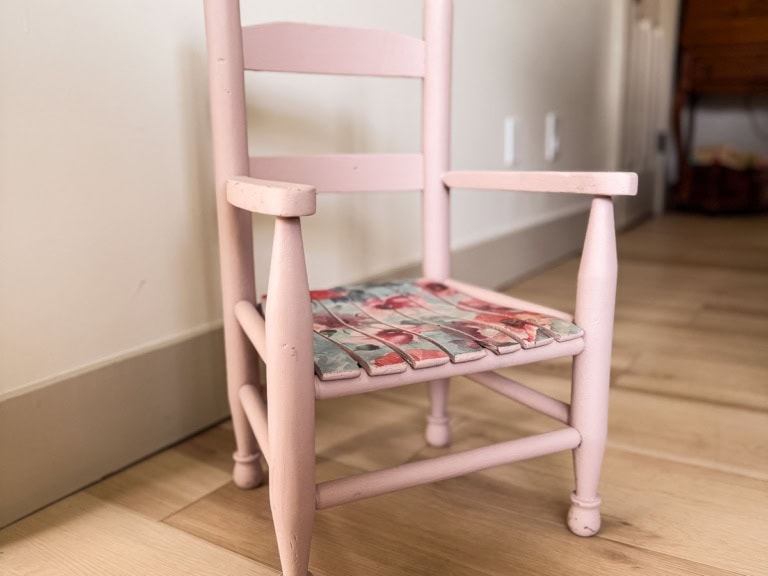 A small, light pink wooden chair with a decorative, multicolored woven seat sits on a light wood floor near an off-white wall.