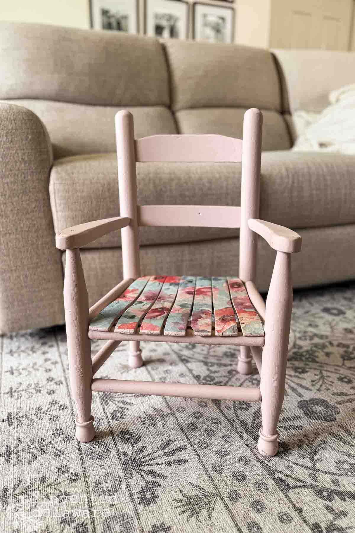 A thrift flip chair makeover: this small pink wooden chair with a floral-patterned woven seat sits on a patterned rug, in front of a beige sofa in a cozy living room.