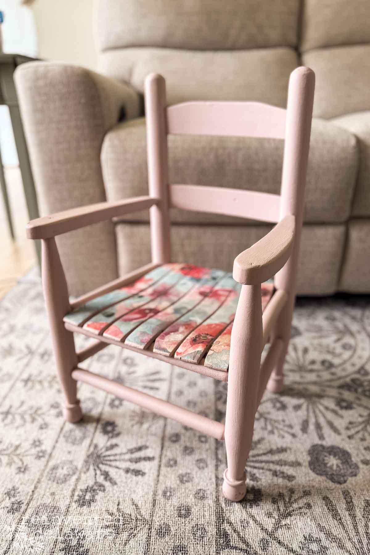 A small, pink wooden chair with a floral cushion—showcasing a charming decoupage chair makeover—sits on a patterned rug in front of a beige sofa.
