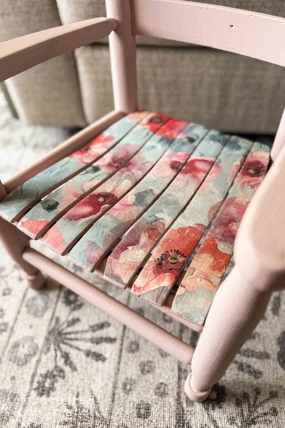 A wooden chair with a decoupage chair makeover, its seat adorned in a colorful floral pattern of red and pink flowers on blue. The chair is painted light pink and sits atop a patterned rug.
