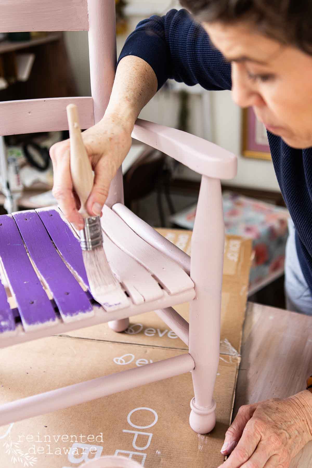 A woman uses a paintbrush to apply light pink paint to an upcycled chair, covering part of the seat that was previously painted purple. The chair, undergoing a colorful makeover, sits on a cardboard surface.