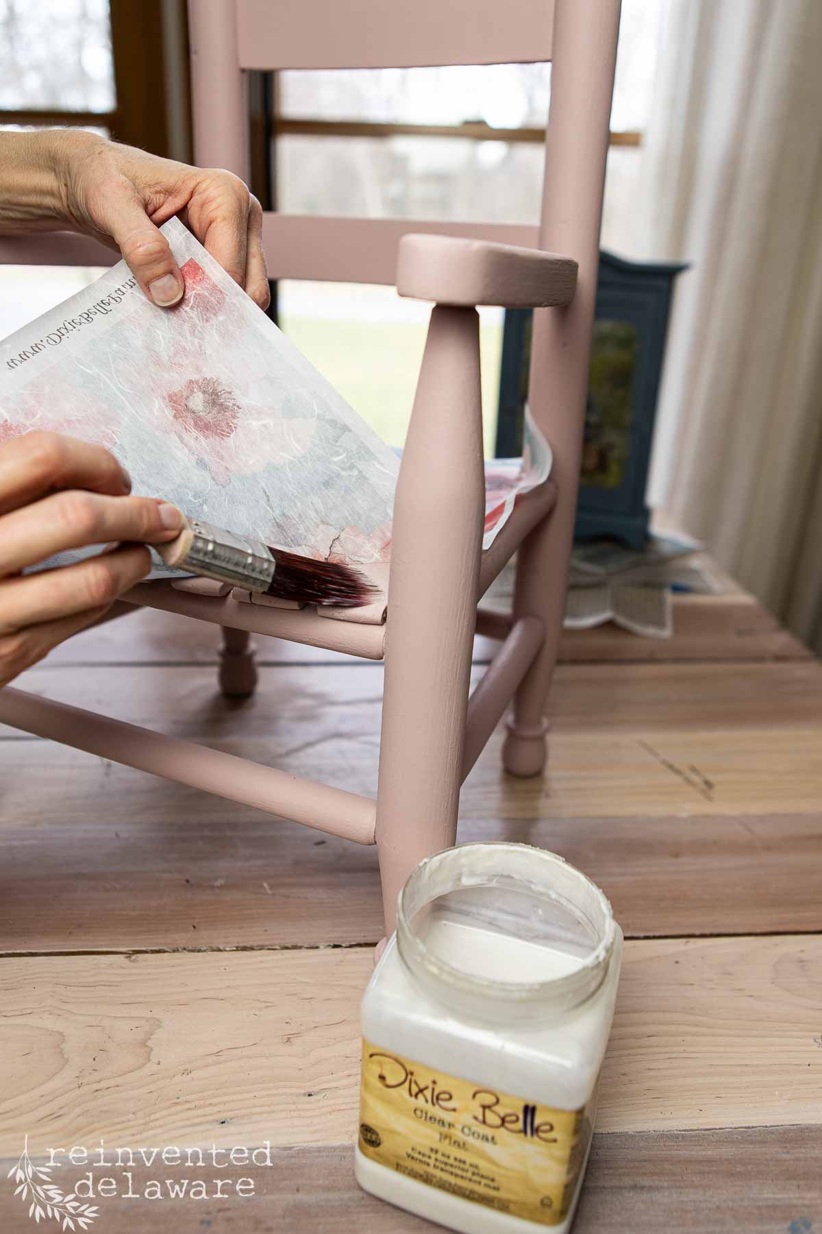 A person uses a paintbrush to apply decoupage glue to a pink wooden chair while holding a floral napkin; an open jar of Dixie Belle Clear Coat sits on the floor nearby.
