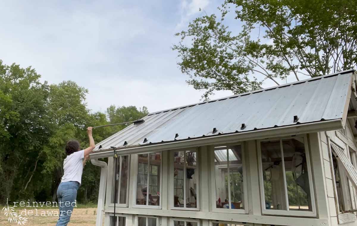 A person stands on the ground, using a long brush to clean the metal roof of a small greenhouse with large windows, surrounded by trees and a cloudy sky.