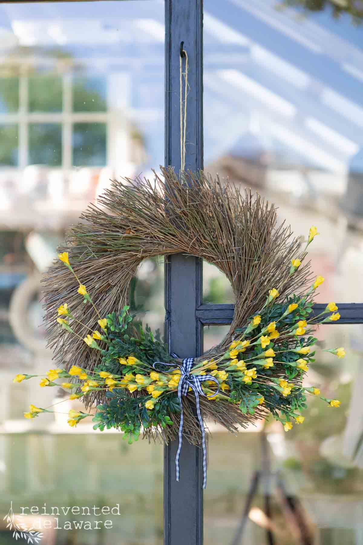 A rustic wreath made of twigs, decorated with small yellow flowers, green leaves, and a blue-and-white checkered ribbon, hanging on a glass door.
