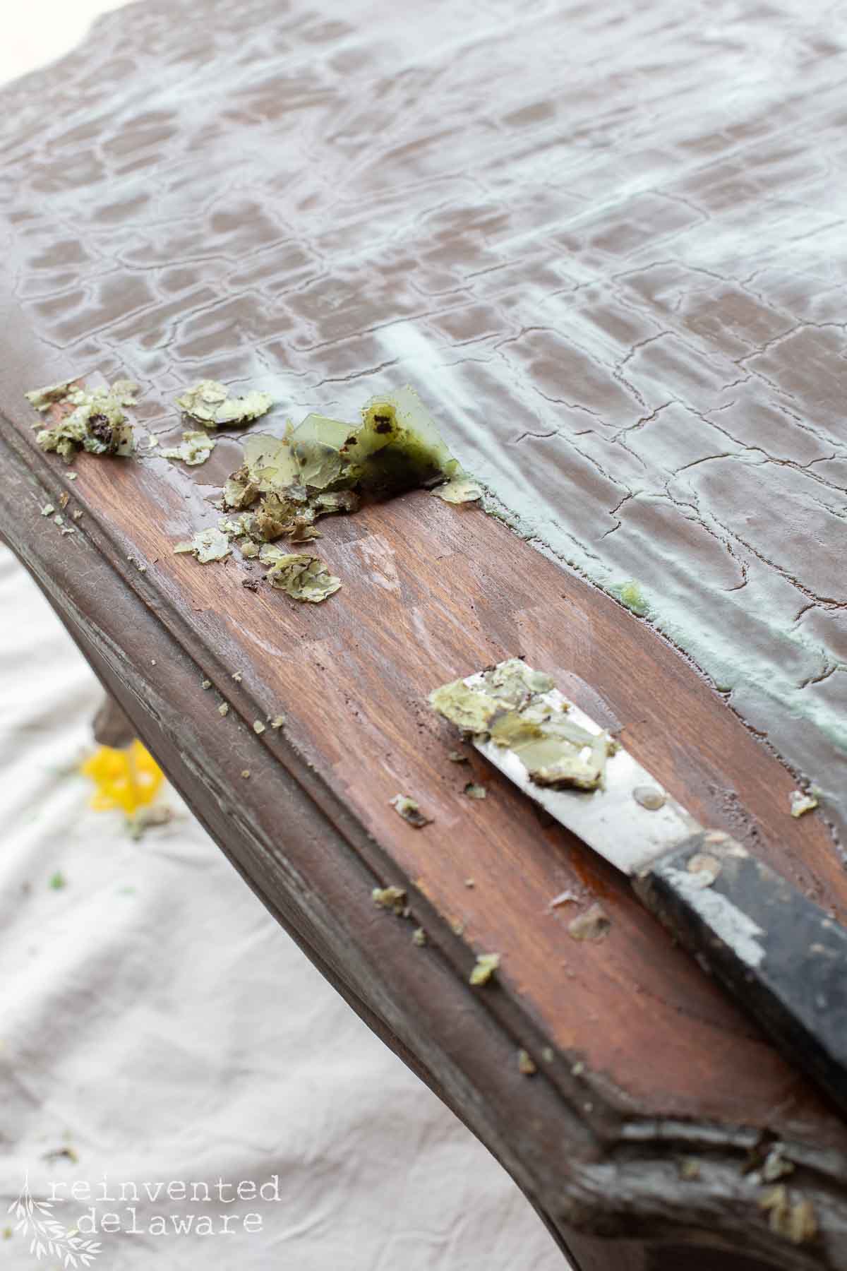 A close-up of a wooden table being stripped of old stain with a metal scraper. Paint chips and residue are visible on the table and scraper, revealing the wood underneath.
