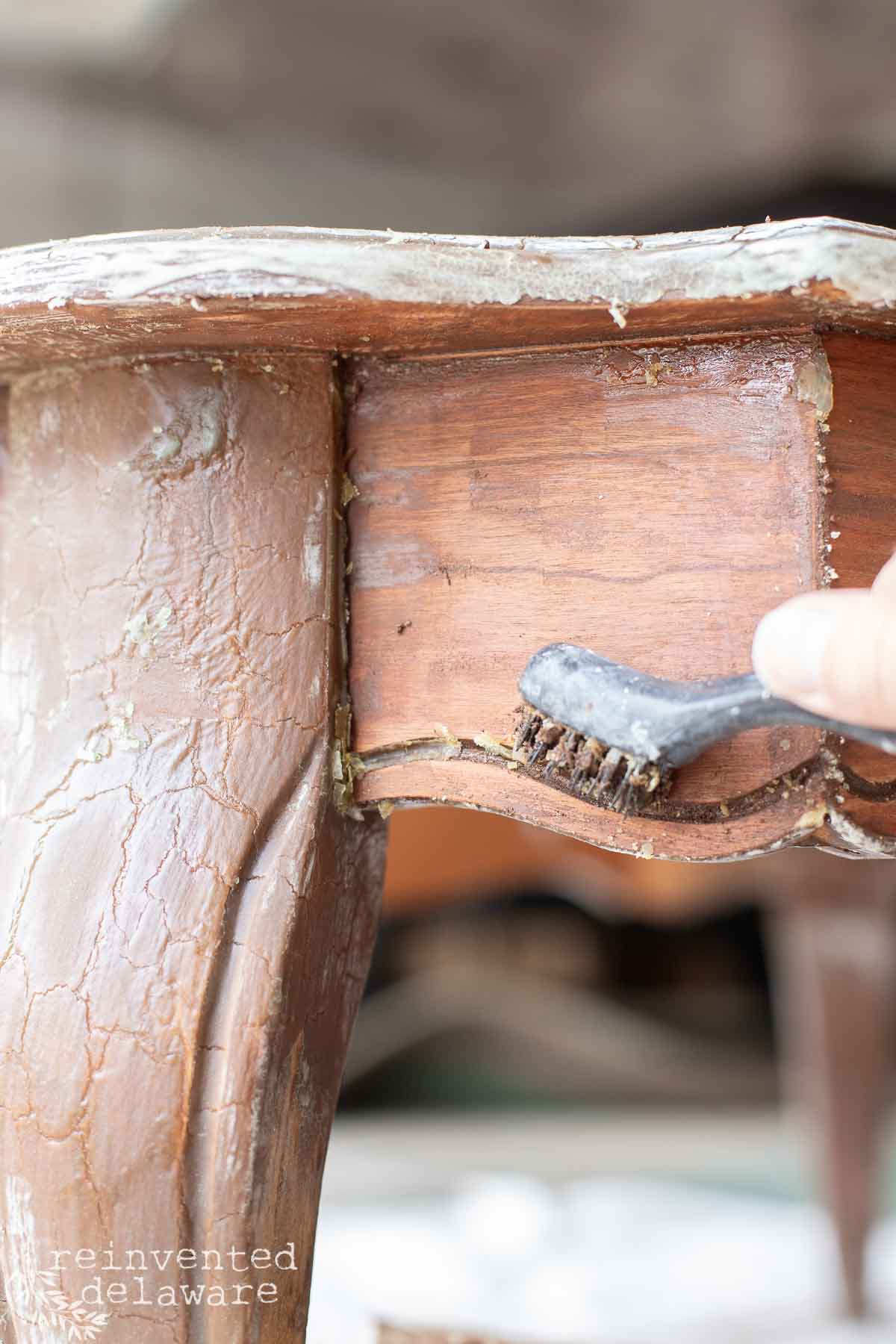 A person uses a small brush to clean or apply a substance to the joint of a wooden, antique table leg with chipped old stain finish.