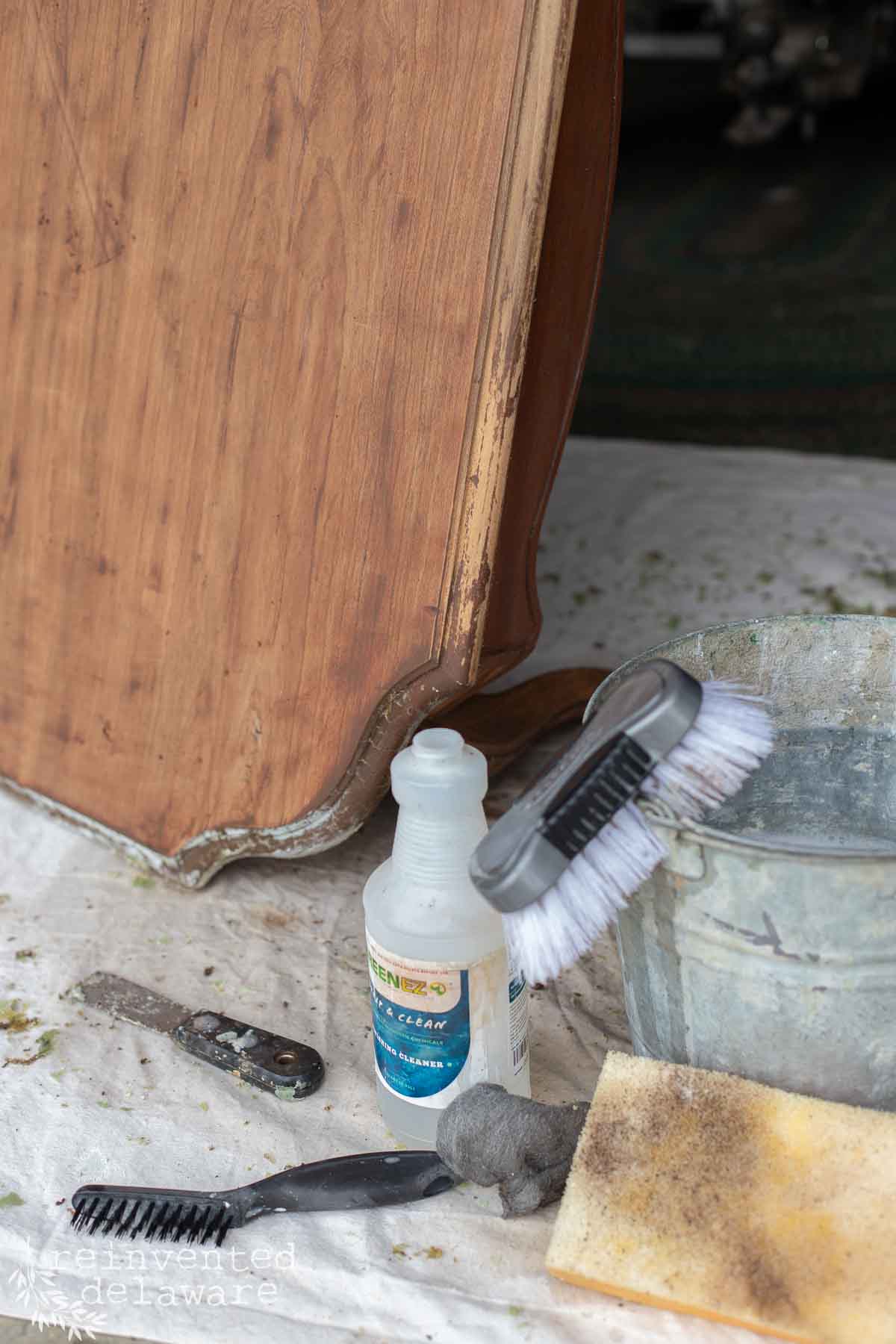 A close-up of furniture cleaning supplies, including brushes, a sponge, steel wool, a cleaning solution, and a metal bucket, next to a wooden cabinet on a drop cloth.