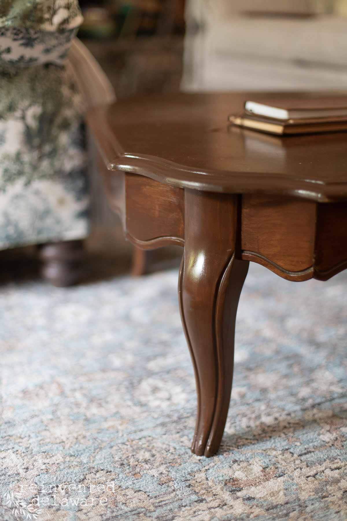 Close-up of a dark wooden coffee table with curved legs, placed on a light patterned rug. A floral-patterned upholstered chair and two stacked books are partially visible in the background.