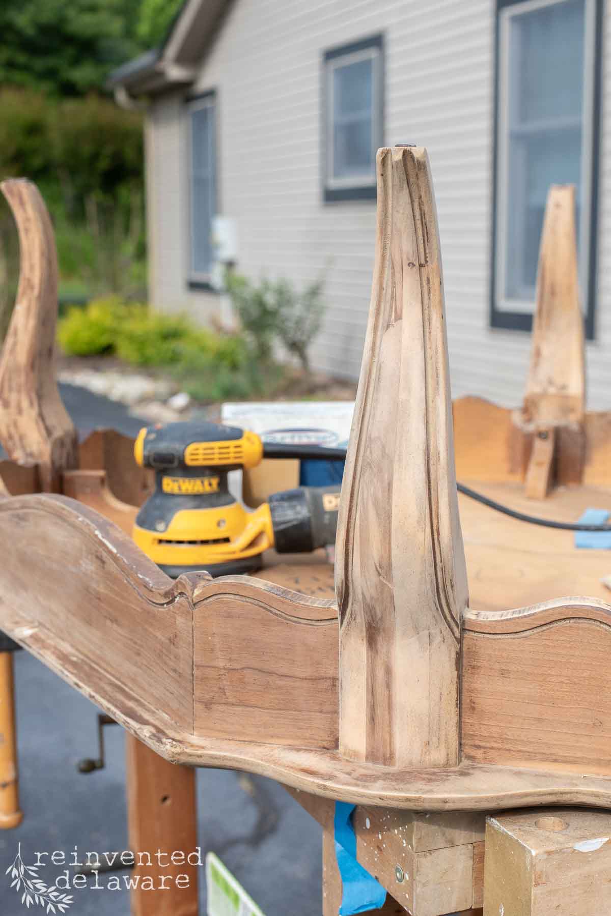 An upside-down wooden table is being sanded outdoors. A power sander rests on the table’s surface, and the table legs show signs of sanding. Greenery is visible in the background.