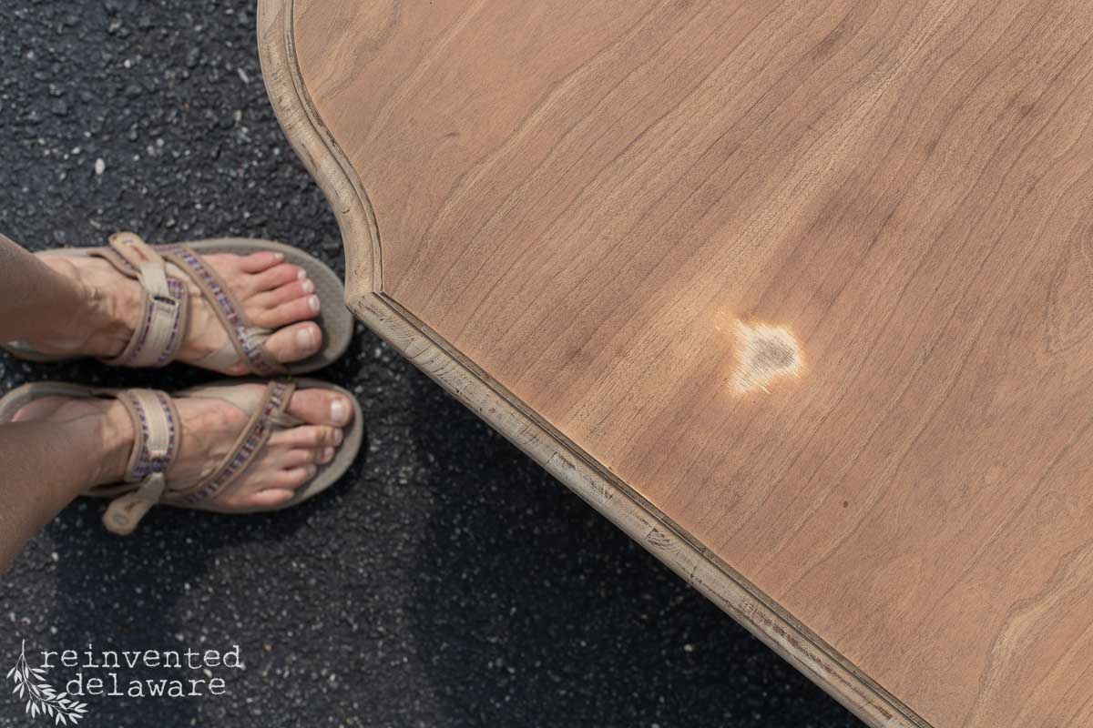 A person wearing sandals stands next to a wooden tabletop with a noticeable burn mark and areas of damaged veneer. The table edge has curved details, and the surface beneath is blacktop. "reinvented delaware" text is in the bottom left corner.