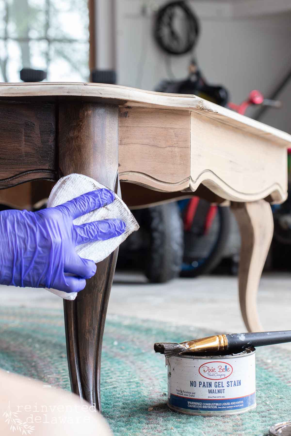 A person wearing a purple glove stains a wooden table leg with a cloth. An open can of walnut gel stain and a paintbrush are on the floor. The background shows a rug, a clock on the wall, and a bicycle.