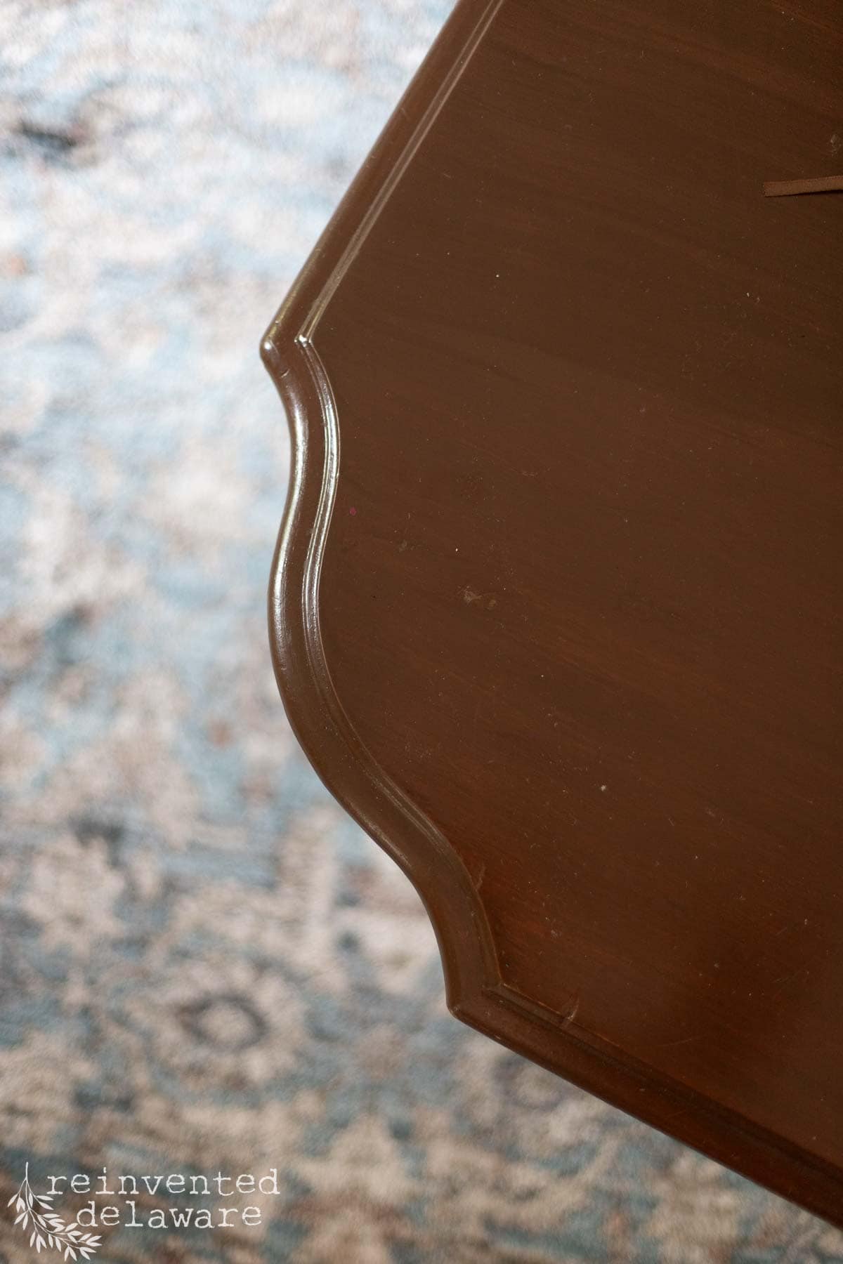 Close-up of a dark wooden coffee table with curved legs, placed on a light patterned rug. A floral-patterned upholstered chair and two stacked books are partially visible in the background.