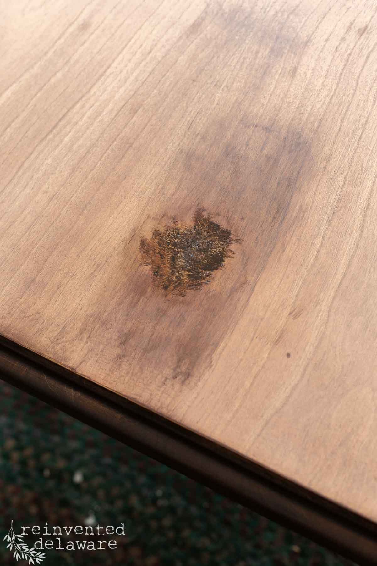 A wooden tabletop with a dark, irregular burn mark in the center and a light wood grain finish, perfect for those looking to repair damaged wood veneer. The "reinvented delaware" logo is in the bottom left corner.