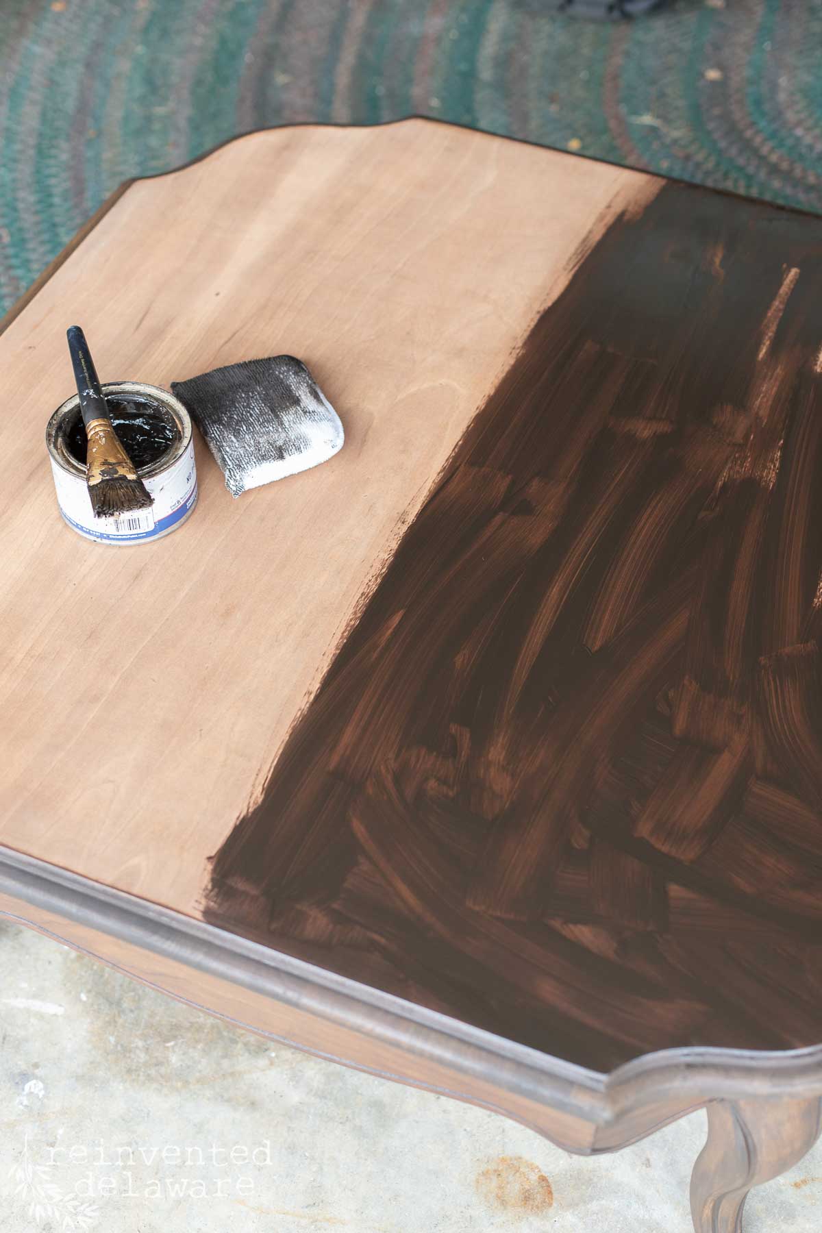 A wooden table being stained; half is bare while the other half is coated dark brown. A paintbrush and an open can of stain rest on the unfinished side.