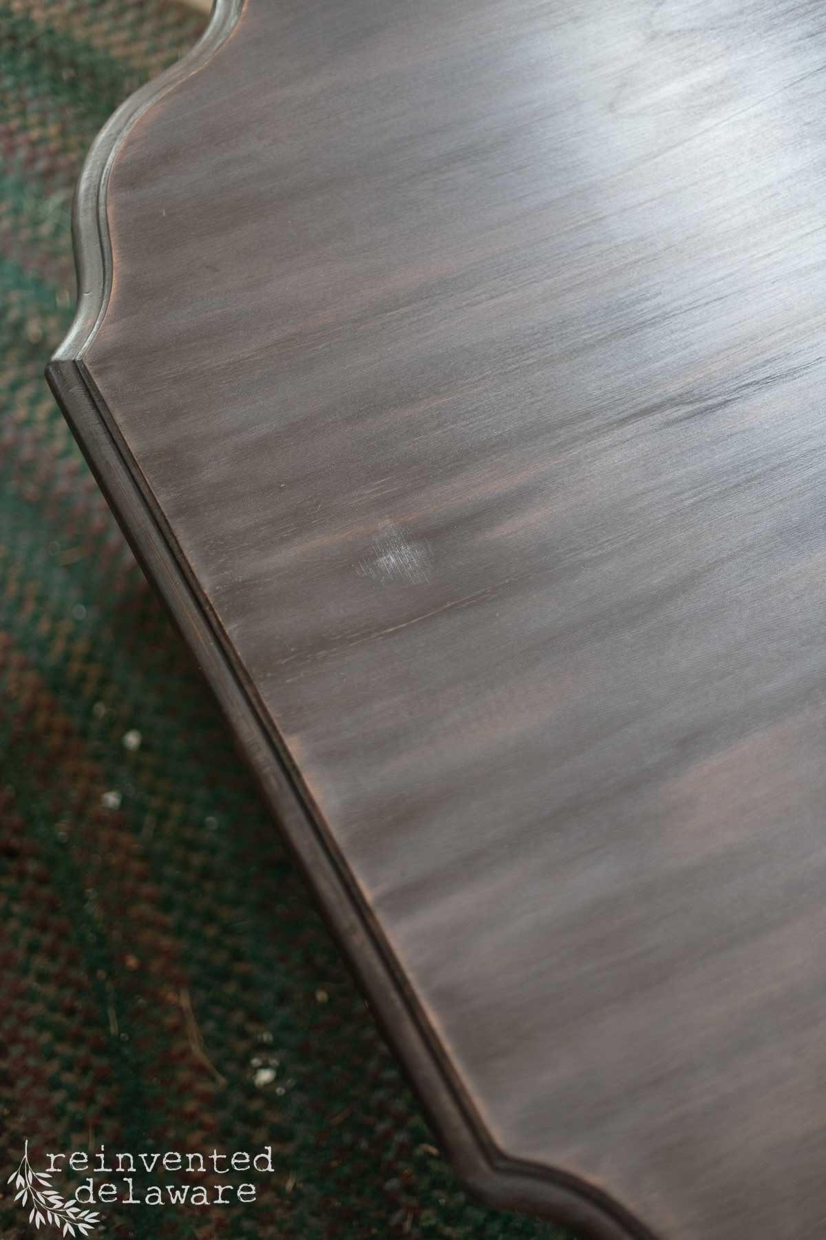 A close-up of a polished wooden tabletop with a beveled edge, showing a small smudge or imperfection on the surface—an ideal candidate for repairing damaged wood veneer. The corner of a green patterned fabric peeks out from underneath. "reinvented delaware" is in the lower left corner.