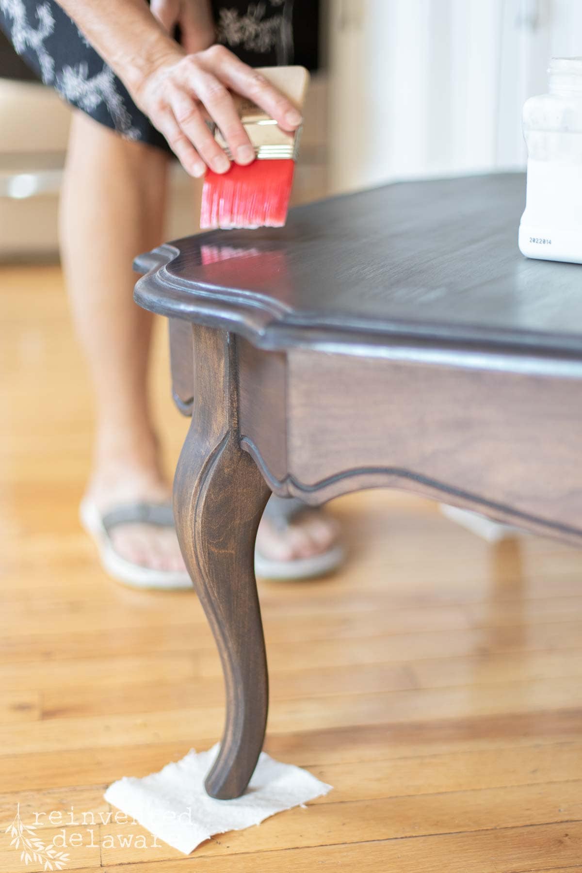 A person paints the edge of a dark wooden table with a red brush, carefully preparing to apply clear coat on the stained wood surface for a smooth, protective finish. The table leg rests on a small piece of white cloth to protect the floor. The person wears sandals and a patterned dress.
