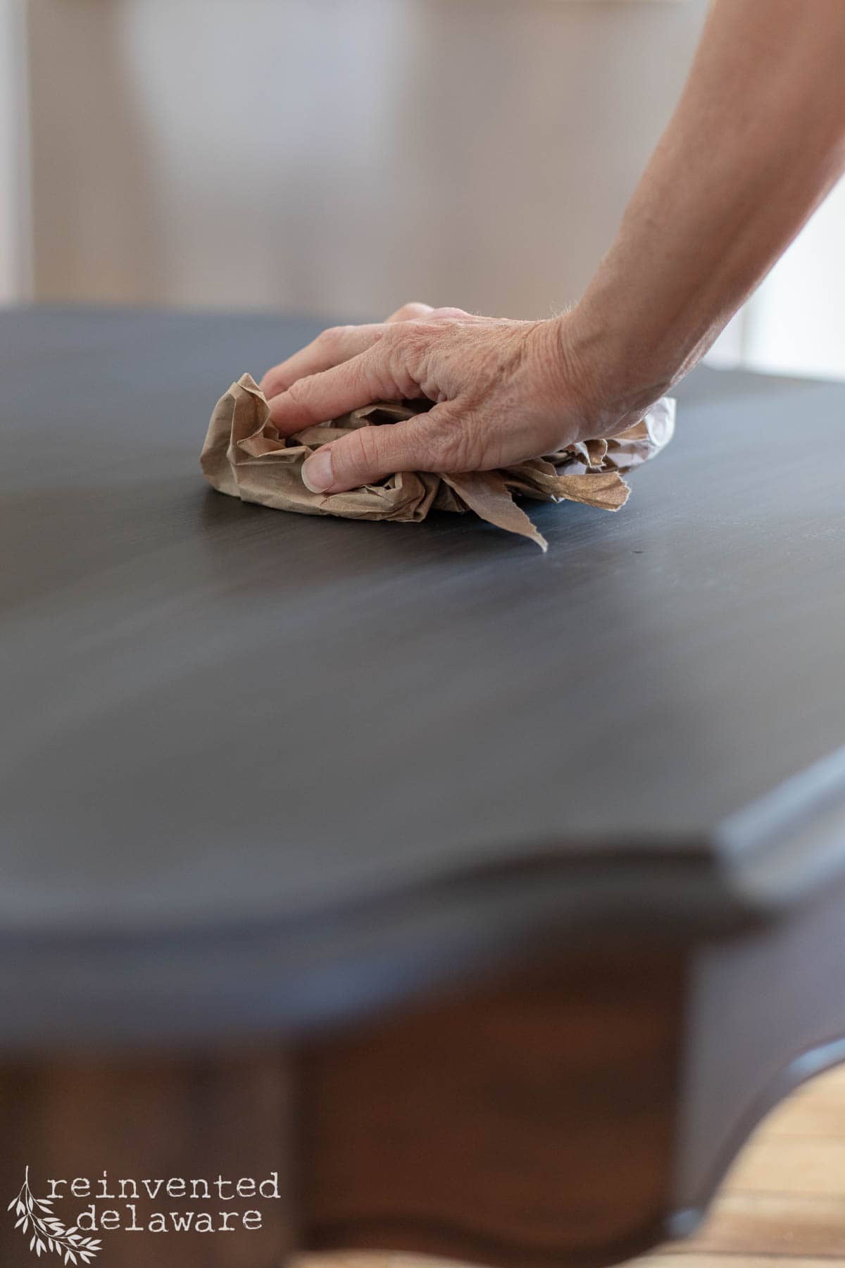 A hand wipes a dark wooden tabletop with a brown cloth. The table has curved edges, and the scene is softly lit. "reinvented delaware" is written in the bottom left corner.