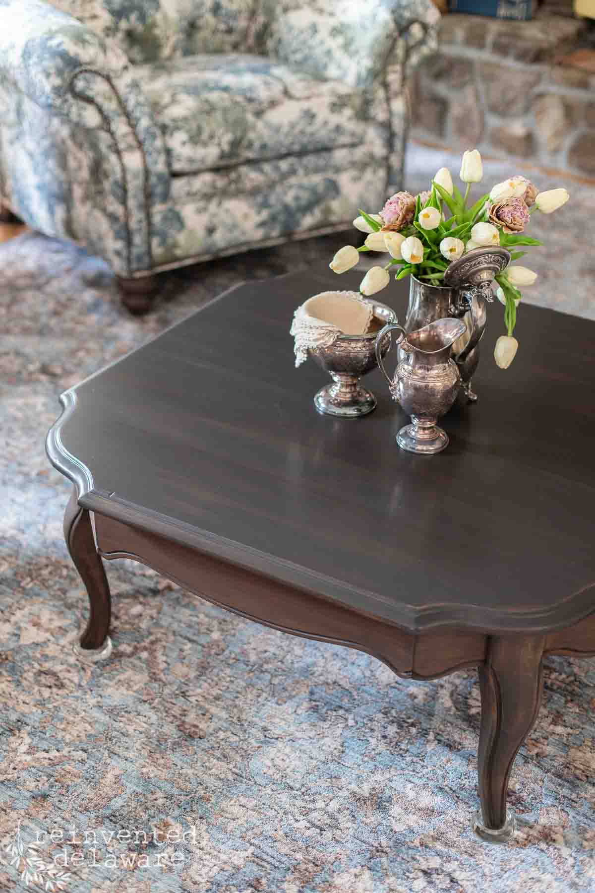 A dark wood coffee table with silver vases, a pitcher, and a bouquet of tulips, set on a patterned rug with a floral armchair and stone fireplace in the background.