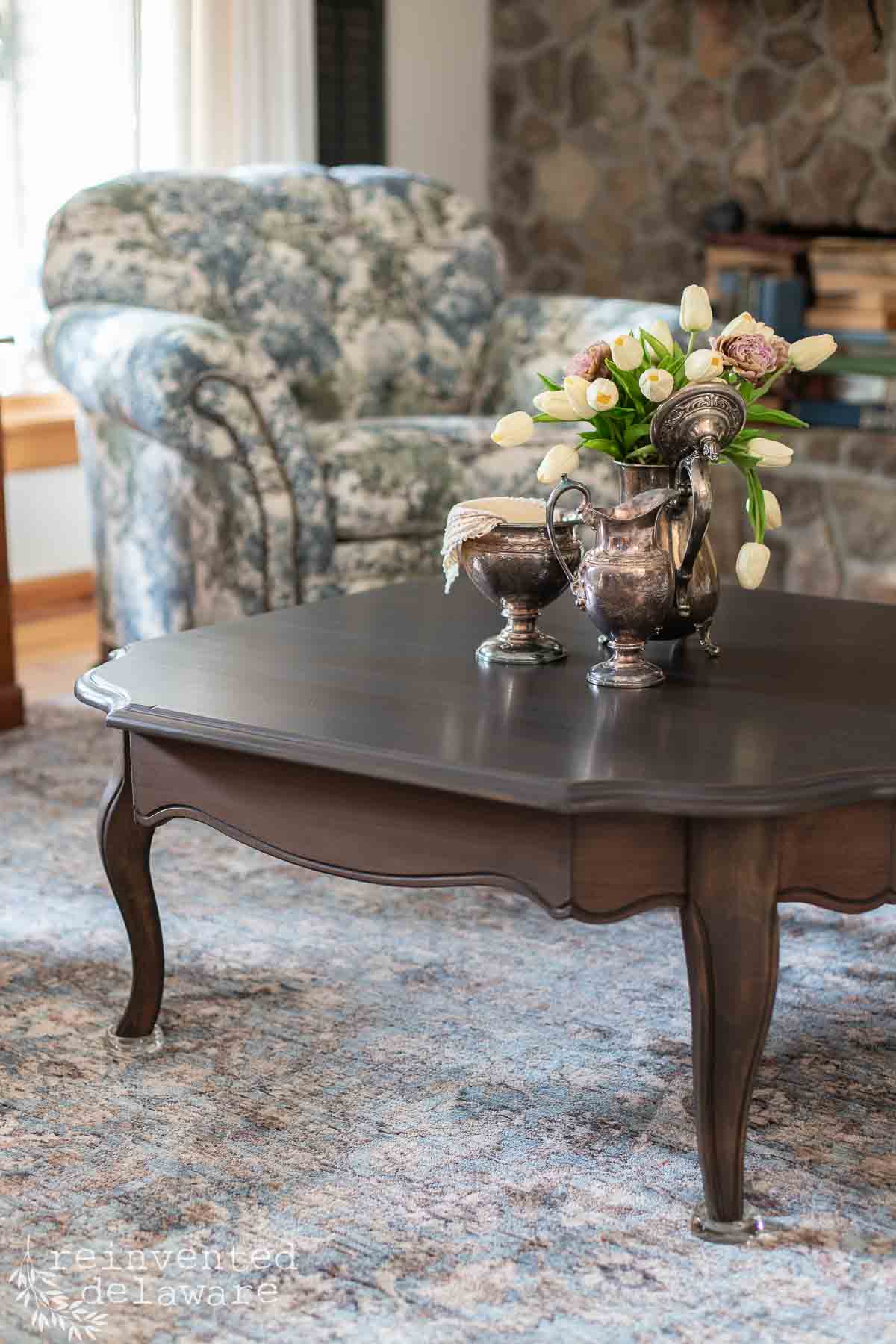 A dark wood coffee table with silver teapots and a floral arrangement sits on a blue patterned rug, in front of a floral armchair and a stone fireplace.