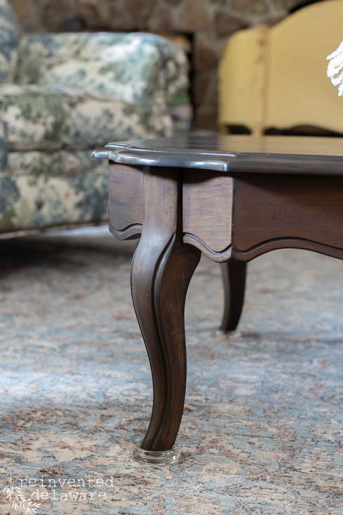 Close-up of a dark wooden coffee table leg with curved, elegant design, shown on a patterned rug. Part of a floral armchair and a yellow sofa are visible in the blurred background.