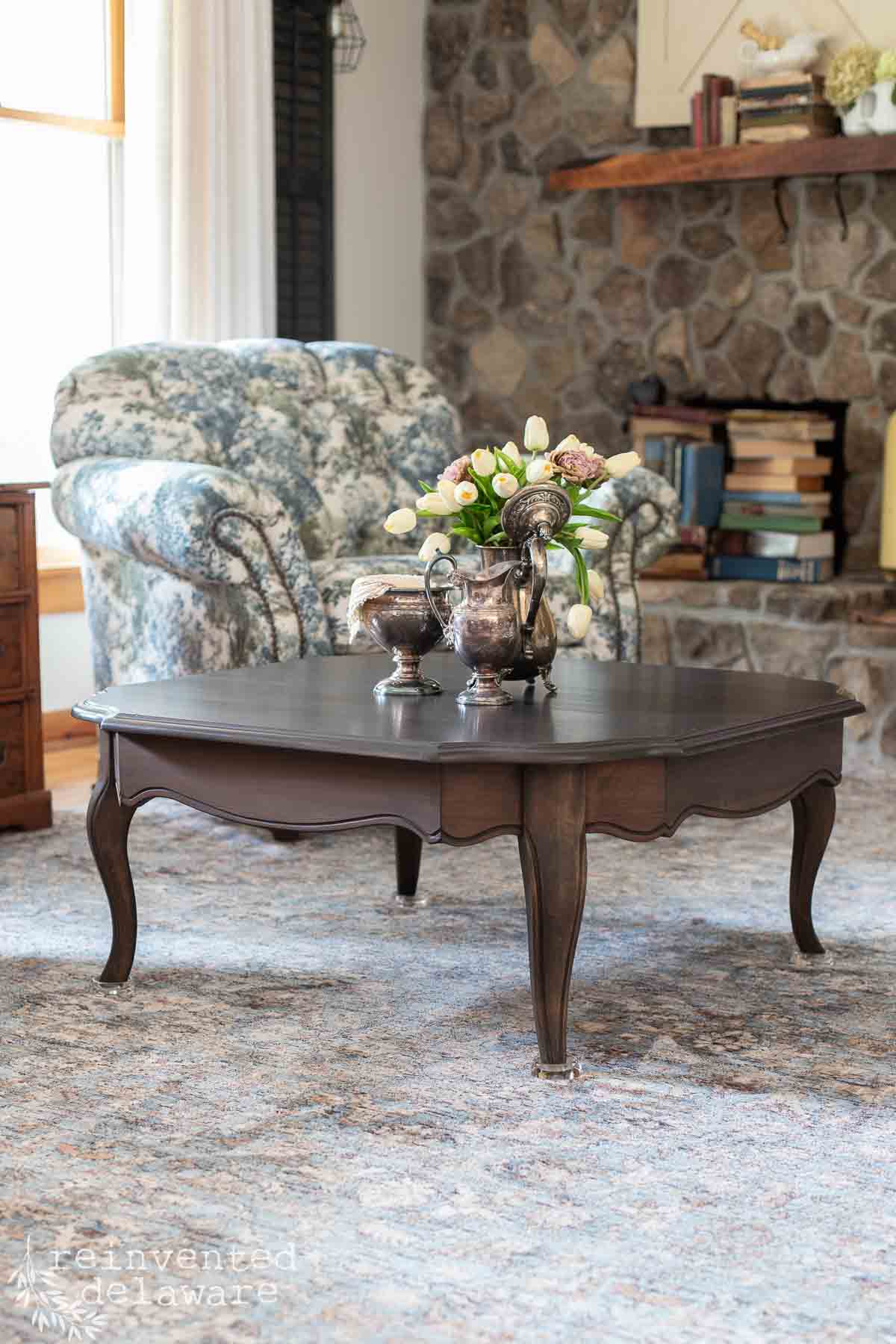 A dark wood coffee table with silver tea set and flowers sits on a blue-patterned rug. Behind it is a floral armchair, a stone fireplace, and shelves with books and decor in a cozy living room.