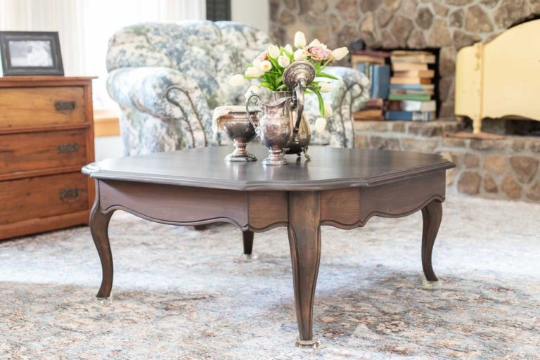 A dark wood coffee table with silver teapots and a flower arrangement sits on a patterned rug in a cozy living room with a floral armchair, stone fireplace, and wooden chest.