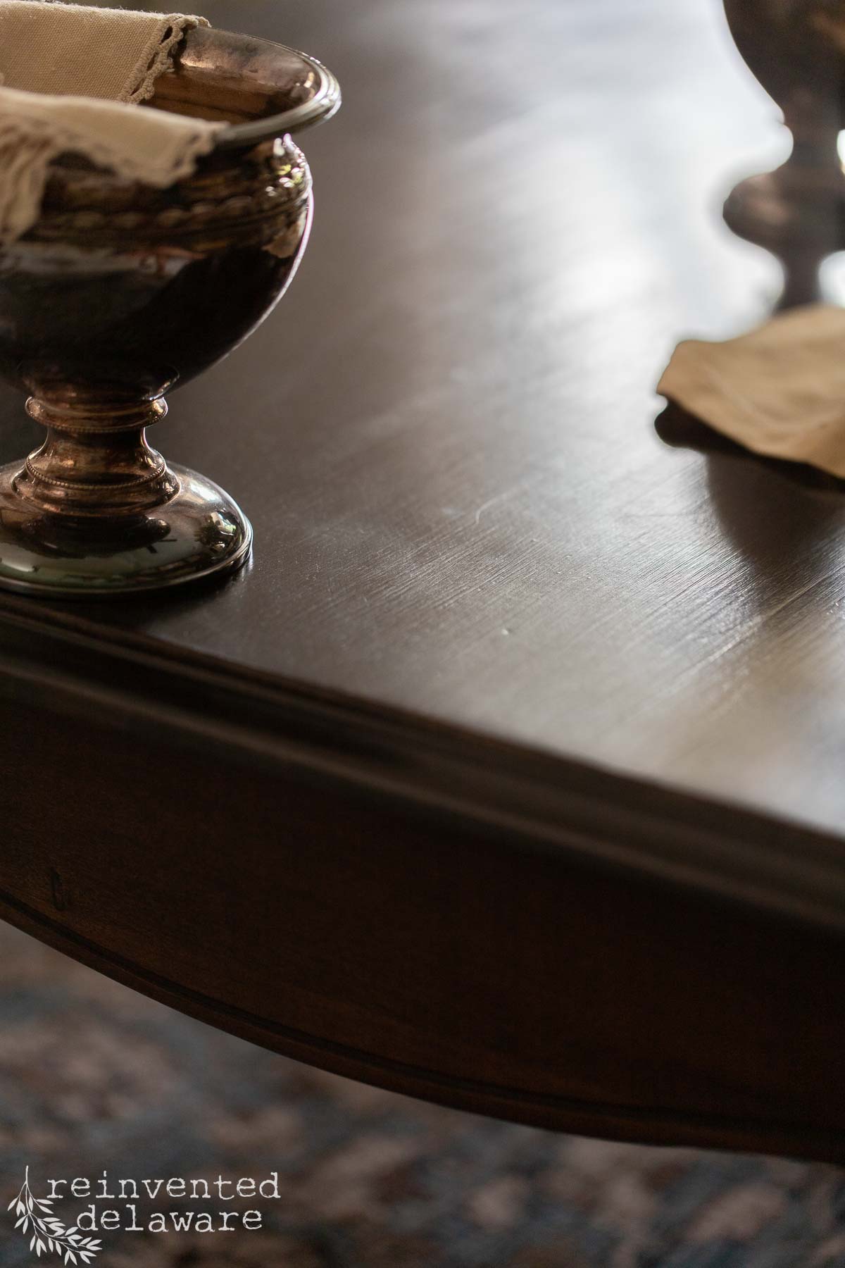 A close-up of a dark wooden table’s edge with a shiny goblet and napkin on top. The table surface reflects light. "Reinvented Delaware" is written at the lower left corner.