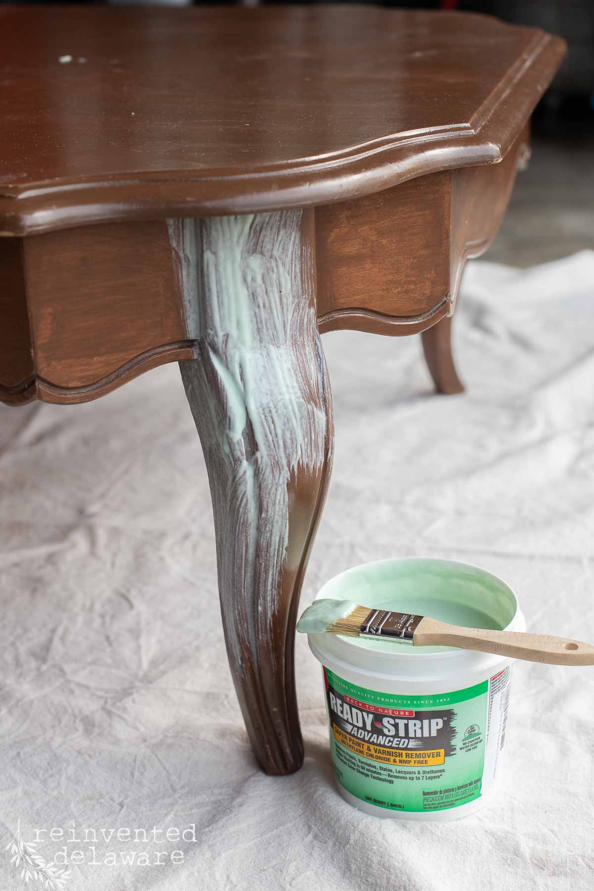 A wooden table leg with paint stripper being applied, a brush resting on an open tub of Ready-Strip on a drop cloth beneath the table.