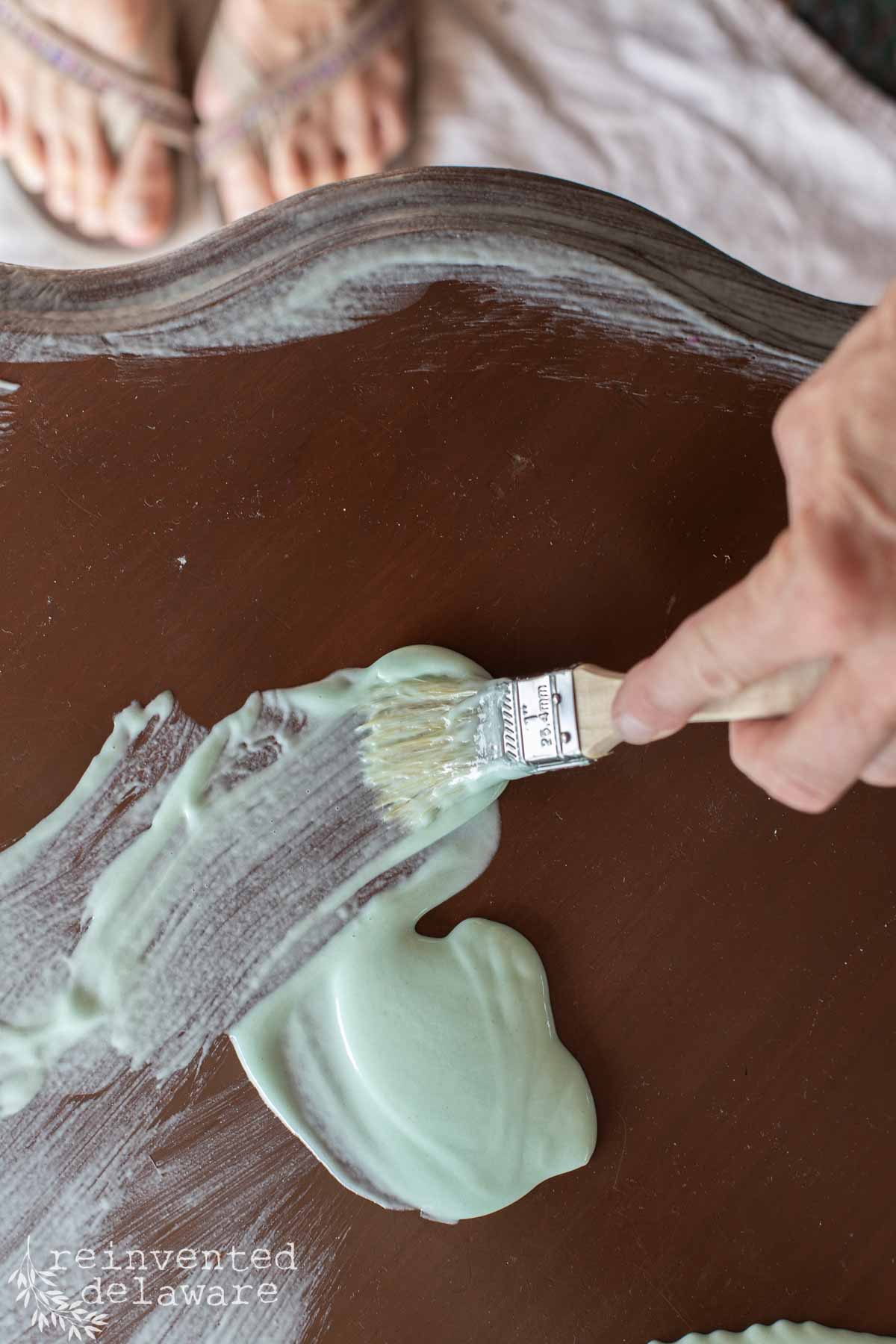 A hand uses a paintbrush to apply pale green paint stripper onto a dark wooden surface, with a pair of bare feet visible at the top of the image.